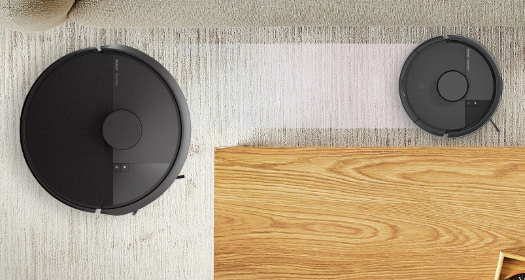 L'immagine dall'alto mostra due robot aspirapolvere iRobot Roomba di colore nero opaco su un pavimento con tappeto chiaro e parquet in legno.