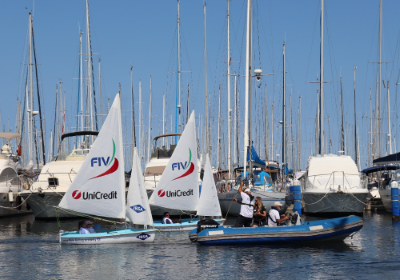 L'immagine mostra un momento di attività in mare all'interno di un porto turistico, sotto un cielo azzurro e limpido, con le barche a vela bianche da regata inclusiva (classe Hansa 303) 