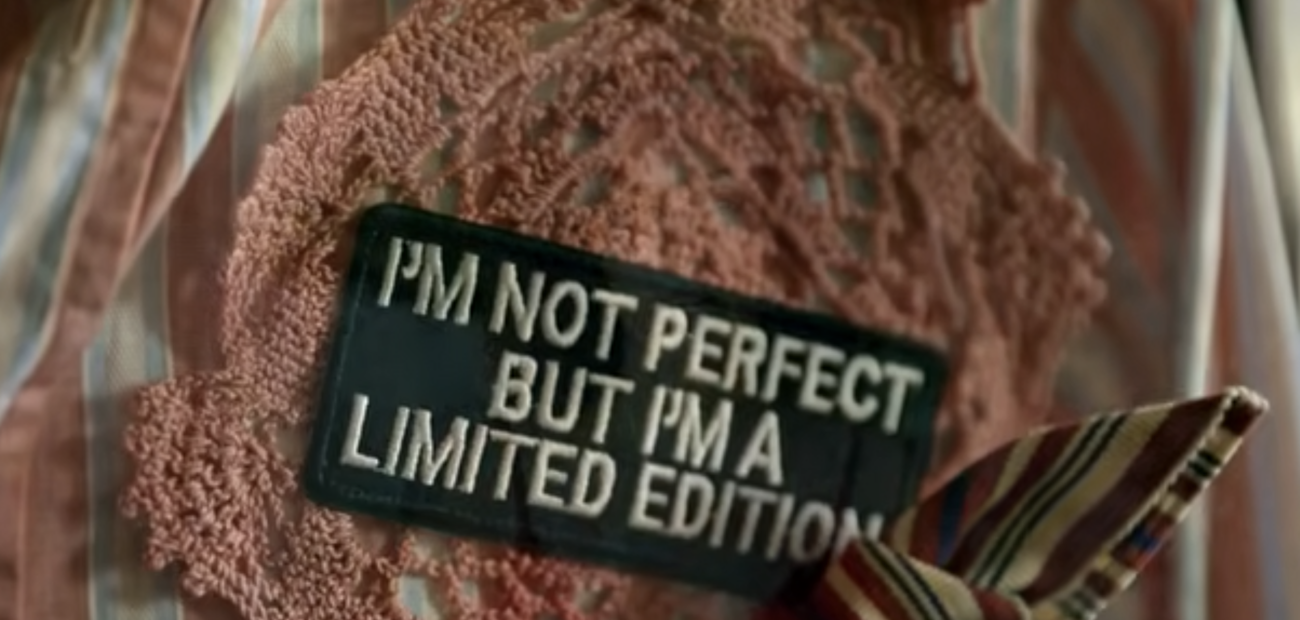 Dettaglio in primo piano una etichetta rettangolare nera a con lettere bianche ricamate in rilievo che riportano la scritta in inglese: «I'M NOT PERFECT BUT I'M A LIMITED EDITION» (Non sono perfetto ma sono un'edizione limitata). L’etichetta è appoggiata sopra un inserto decorativo rosa dalla trama traforata.