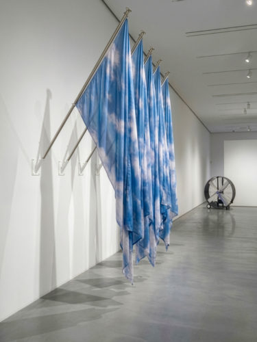 Paolo Puddu – Personne (2026). Fissate al muro tramite aste metalliche inclinate a quarantacinque gradi, sono appese quattro grandi bandiere in tessuto. La stoffa di ciascuna bandiera è stampata con l'immagine fotografica di un cielo azzurro solcato da nuvole bianche. Le bandiere ricadono in ampie pieghe sinuose. Sullo sfondo un grande ventilatore industriale circolare in metallo. L'opera materializza il cielo e il vento dentro lo spazio chiuso del museo. 