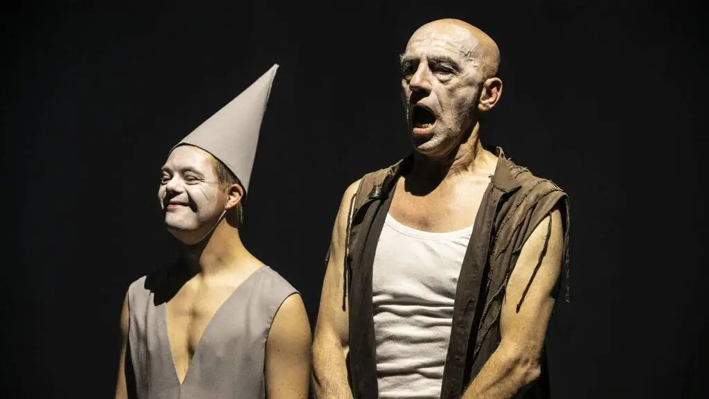 Due attori in scena durante La Tempesta, immortalati su uno sfondo scuro e illuminati da una forte luce di taglio con un effetto di chiaro-scuro che mette in risalto l’espressività dei volti. L’attore a sinistra indossa uno smanicato con scollo a V e un cappello a cono grigio. Ha il viso truccato di bianco e sorride. L’attore a destra: È un uomo calvo con canottiera bianca e un gilet marrone. Ha il viso parzialmente sbiancato dal trucco ed ha la bocca spalancata.